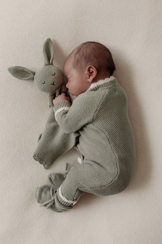 Sleeping newborn baby wearing sage green knit romper cuddling sage green bunny comforter