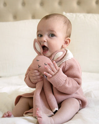 Baby sitting up on a bed cuddling a pink bunny comforter
