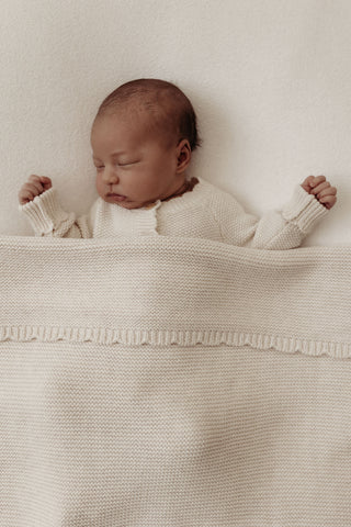 Sleeping baby wearing beige knitted romper and tucked in under a beige knitted blanket with a scalloped edge. 
