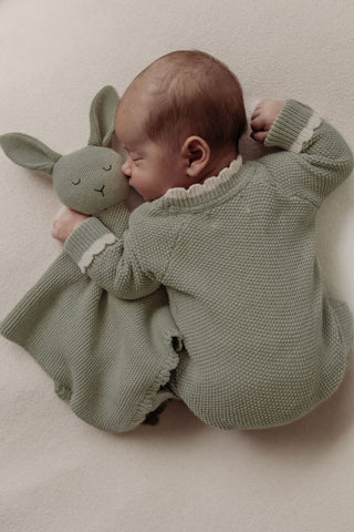 Sleeping baby wearing sage knitted romper cuddling sage green bunny comforter