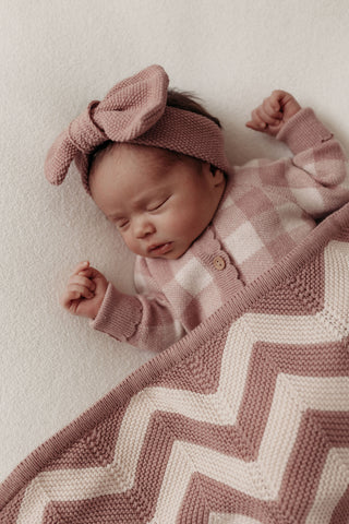 sleeping baby wearing knitted pink bow headband and checkered knit romper, with a pink and cream chevron stripe blanket 