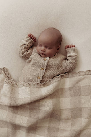 Sleeping newborn wearing a grey knitted romper covered with a grey gingham check blanket with scalloped edges