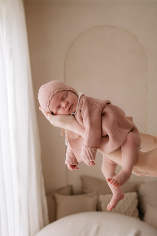 Sleeping baby being lifted up wearing a pink knitted bonnet and cardigan