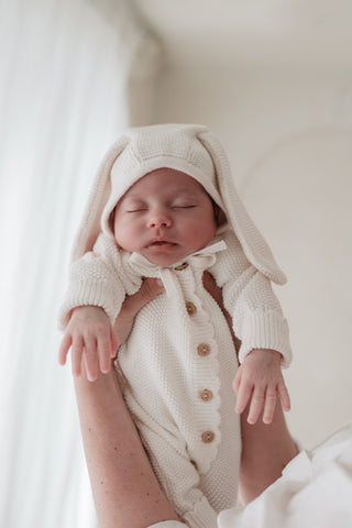 Newborn baby being lifted up wearing a knitted white bunny ear bonnet and matching romper