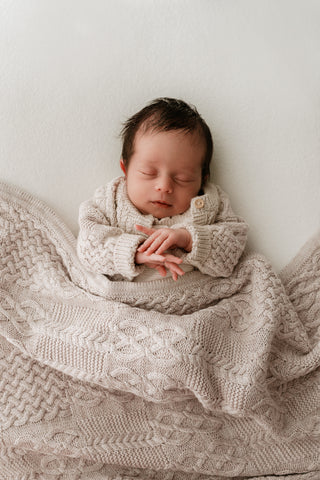 Baby sleeping under a beige cable knit blanket wearing a matching cardigan