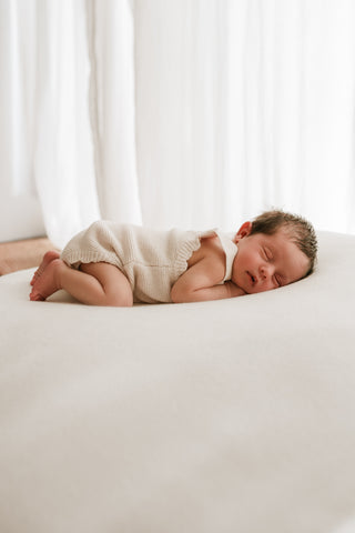 Sleeping baby lying on a beanbag with a sheer curtain behind wearing a neutral knitted romper