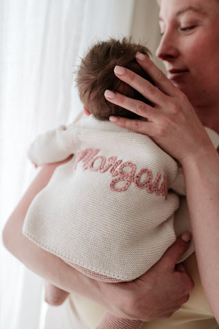 Mother holding baby in her arms, baby is wearing an embroidered cardigan with the name 'Margaux' on the back