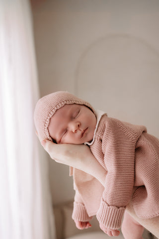 Close up of a sleeping baby being lifted up wearing a pink knitted bonnet and cardigan