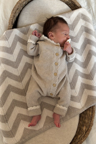 Baby in basket with dummy wearing a knitted babygrow romper, lying on a chevron stripe knitted blanket
