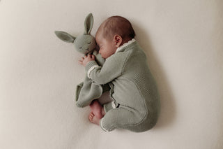 Sleeping baby lying on their side wearing a sage green knitted romper and cuddling a sage green knitted bunny comforter