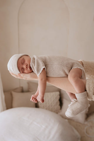 Sleeping baby in mothers arms wearing neutral knitted romper, hat, and booties.