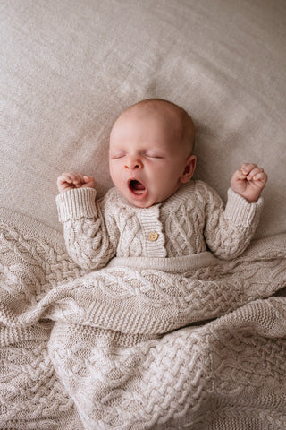 Yawning baby wearing neutral cable knit romper with a matching blanket