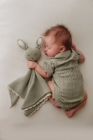 Sleeping baby wearing a sage green knitted bodysuit holding a sage green bunny comforter with a cream scalloped edge