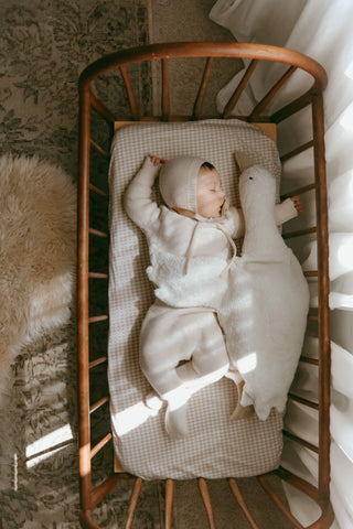 Sleeping baby in cot with goose, wearing knitted bonnet, jumper and trousers, lying on a gingham sheet