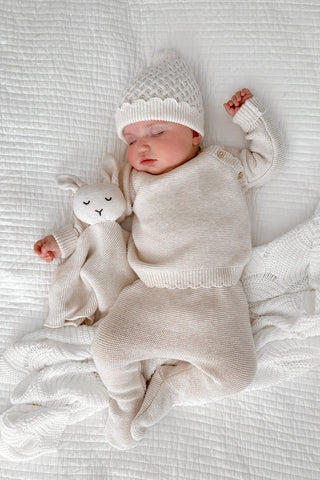 Sleeping baby on white bedding cuddling bunny comforter wearing a beige knitted baby hat, jumper and trousers