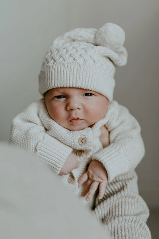 Newborn baby wearing cable knit pom pom hat and knitted romper being held up
