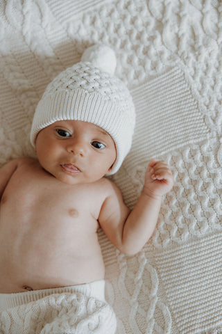 Baby lying on cable knit blanket wearing knitted pom pom hat