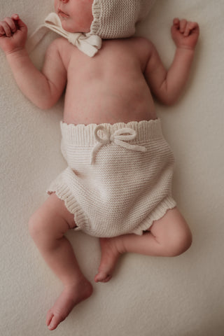 Baby wearing knitted neutral bloomers with bow and scalloped edge