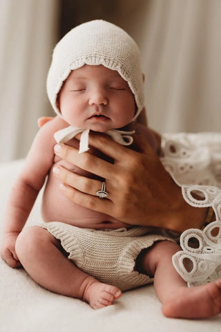 Sleeping newborn baby sitting up being held by mum, wearing a knitted bonnet and bloomers