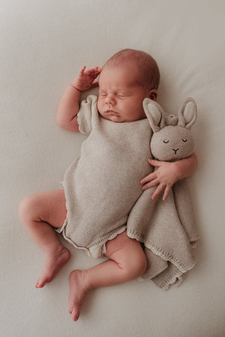 Baby wearing a grey knitted bodysuit with a cream scalloped edge and cuddling a matching grey knitted Isla bunny comforter with a cream scalloped edge.