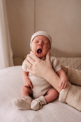 Yawning newborn baby sitting up supported by mother's hands, wearing neutral knitted hat, romper, and booties