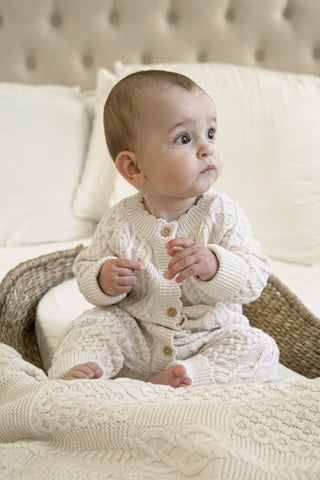 Baby sitting on a bed in a basket wearing cable knit romper