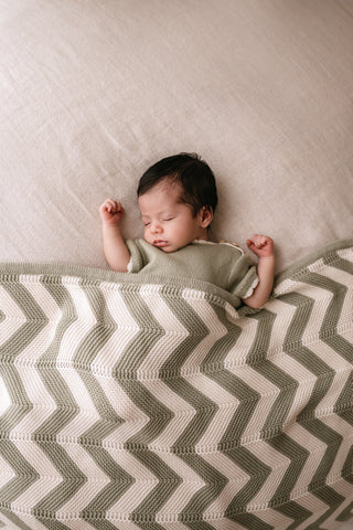Baby wearing a sage knit romper lying under a sage and cream chevron stripe knitted blanket