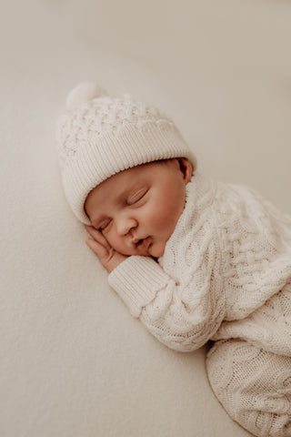 Sleeping baby lying on their front wearing cream cable knit romper and hat