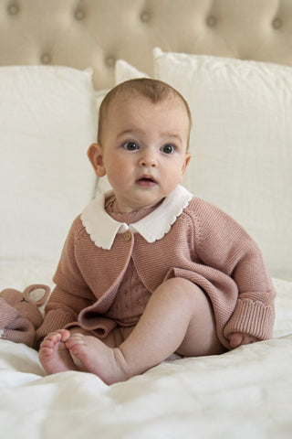 Baby sitting on a bed wearing a pink knitted cardigan with white collar