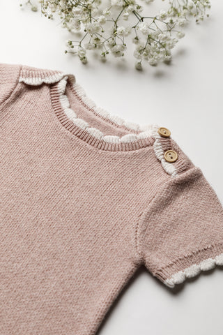 Close up of a knitted pink bodysuit neckline with button shoulder fastening and a cream scalloped trim
